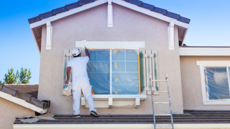 A painter in white attire prepping a residential house for exterior painting, covering windows with plastic sheeting to protect them from paint splatters.