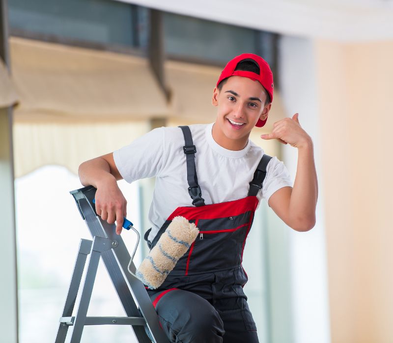 Young painter in overalls and a red cap giving a thumbs up while holding a paint roller, ready to start an interior painting job.