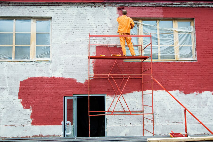 A person from a painting company in an orange outfit painting the wall of a building red while standing on a scaffold.