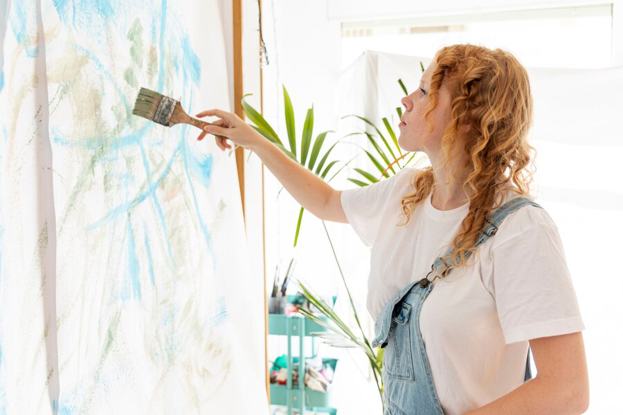 An artist with curly hair attentively adding brush strokes to a large canvas in a bright, plant-filled studio, specializing in painting services.