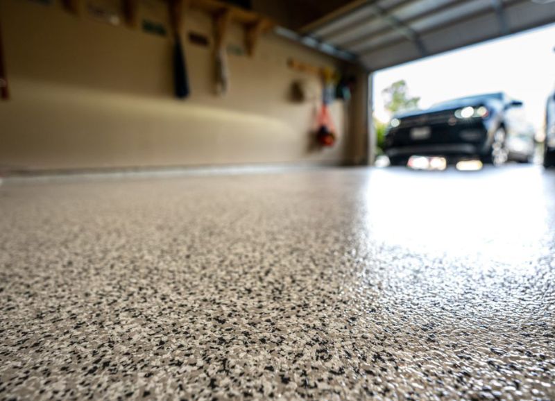 A tidy garage interior with a close-up of a clean speckled floor, featuring recently completed drywall repair, leading towards a parked car in the background.