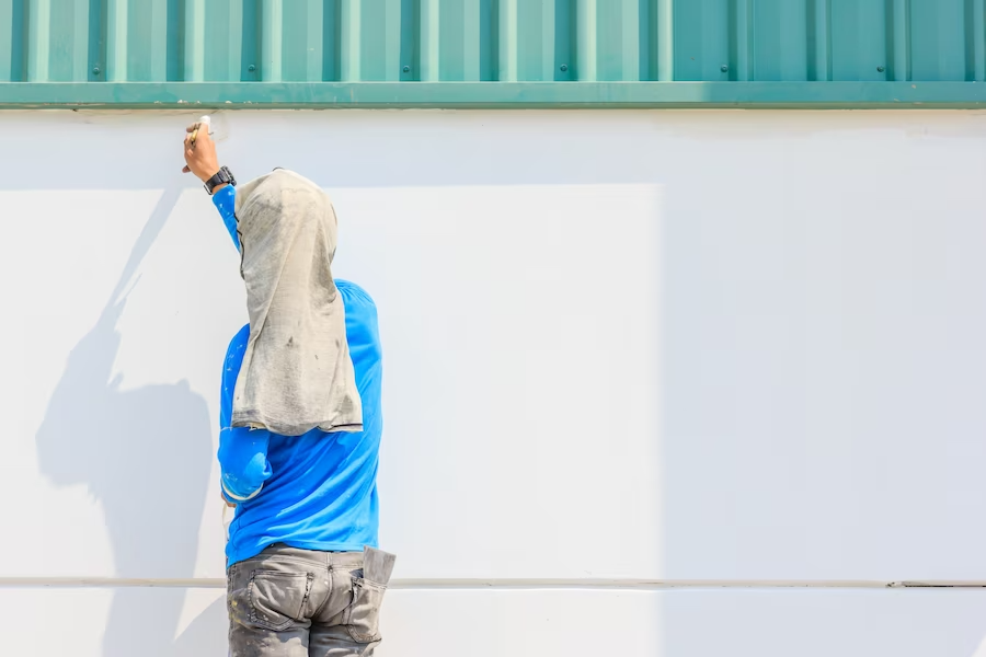 Worker in a blue shirt and gray pants applying paint to a white wall under a green metal roof for a painting company.