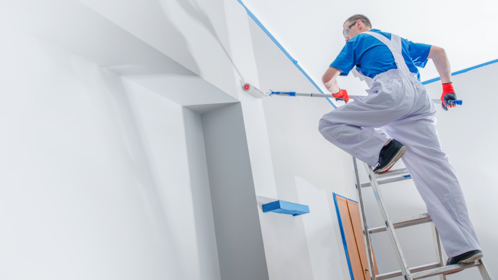 A painter from a professional Painting Company in white overalls and blue shirt meticulously applies paint on a wall with a roller while standing on a ladder, focusing on creating a neat edge where the wall meets the