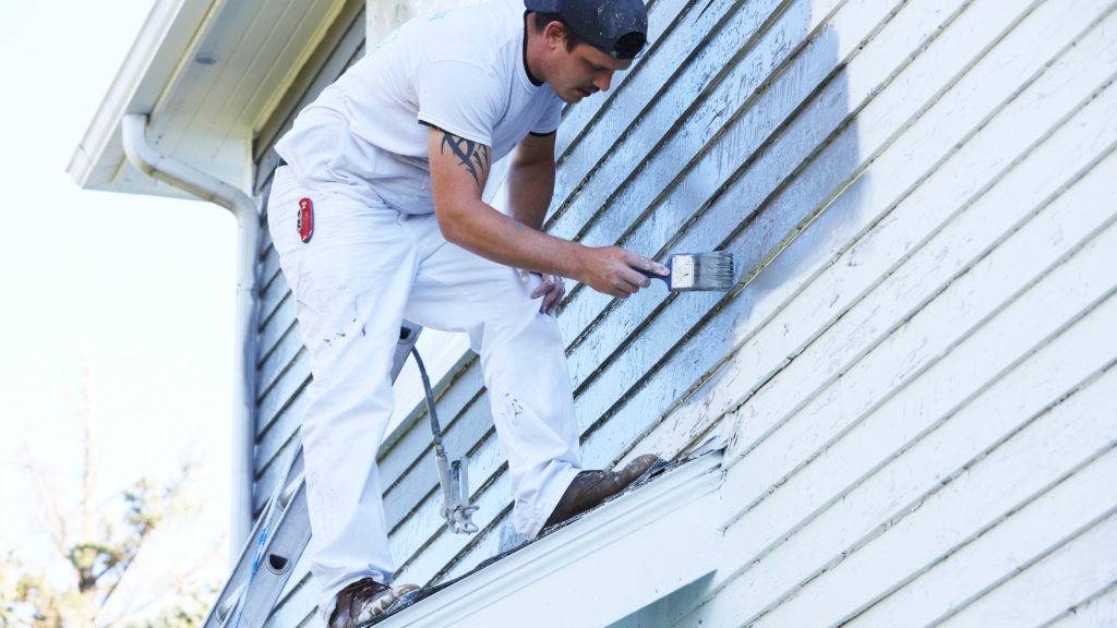 A professional painter in white overalls carefully applying exterior paint to a sidewall while standing on a ladder.