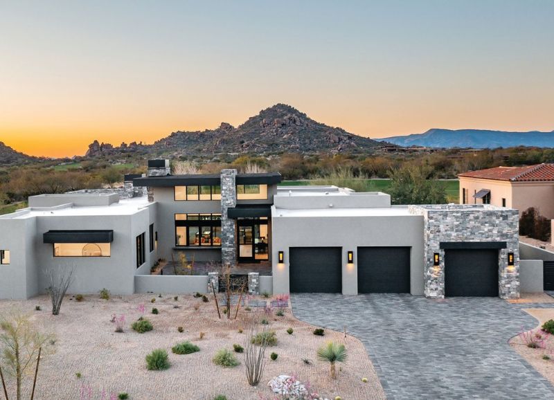 Modern luxury home with desert landscaping at twilight, featuring large windows and a three-car garage against a backdrop of mountain scenery, freshly enhanced by a top-tier painting company.