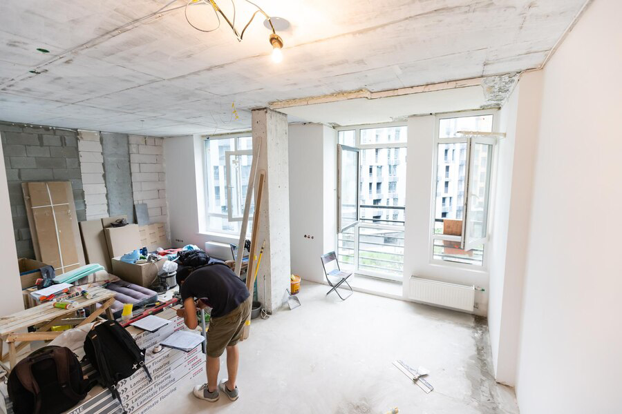 A person working in a spacious, under-construction room with exposed blocks, tools, and building materials scattered around, specializing in interior painting services, with natural light streaming through the windows.