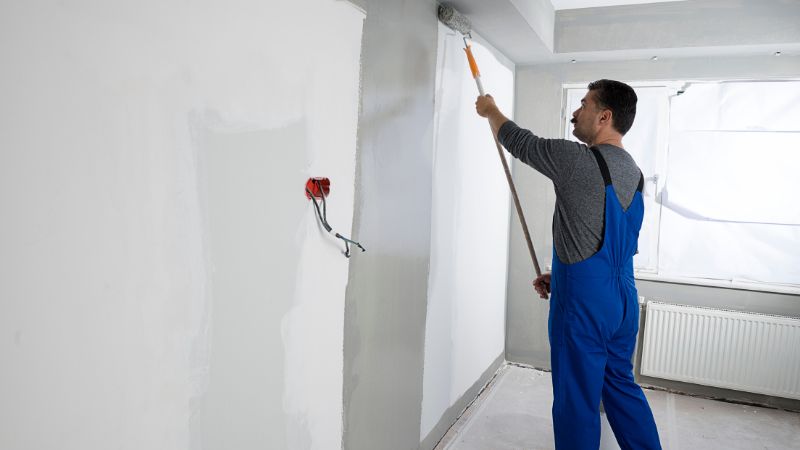 Man in overalls from a painting company applying paint to a wall with a roller brush in a room under renovation.