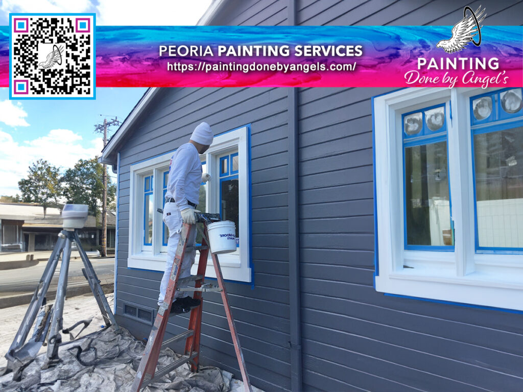 A painter in white coveralls on a ladder applying white paint to trim on a blue building with a sign and QR code advertising Peoria exterior painting services, "painting done by Angel's.