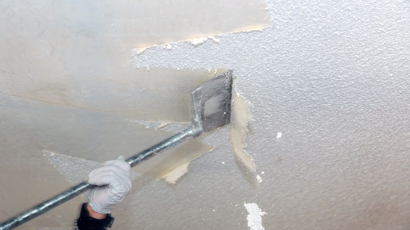 A hand wearing a glove using a putty knife to scrape off peeling paint from a wall corner for drywall repair.