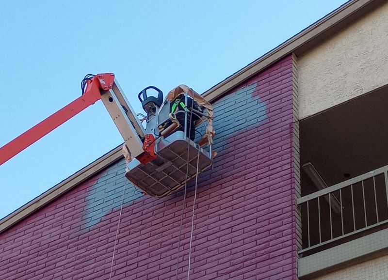Worker in a cherry picker performing maintenance and painting services on a multistory building facade.