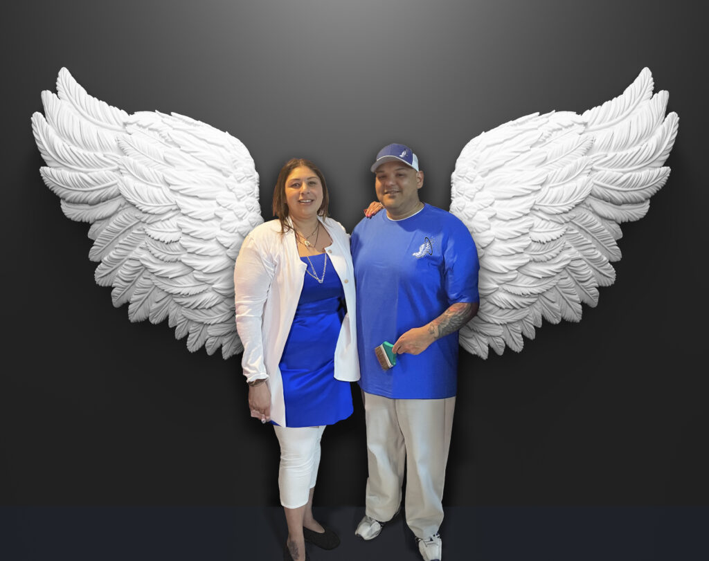 Two smiling individuals standing between a large pair of angel wings painted on the exterior wall, creating the illusion of each having their own set of wings.
