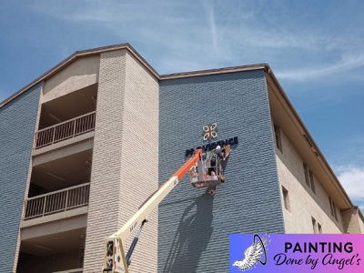 A painting contractor from a Painting Company on a lift refurbishing the exterior wall of a multi-story residential building under a clear blue sky.