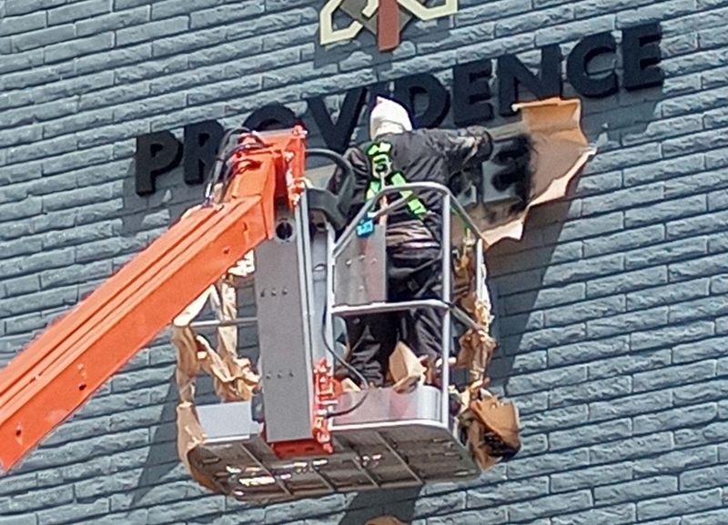 A person in a safety harness is working at height from an orange boom lift, engaged in either installing or repairing a sign on a building facade that reads "providence," or handling exterior painting tasks.
