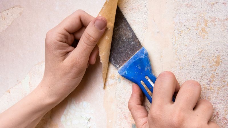 Hands peeling off old wallpaper using a scraper tool during a home renovation, in preparation for interior painting.