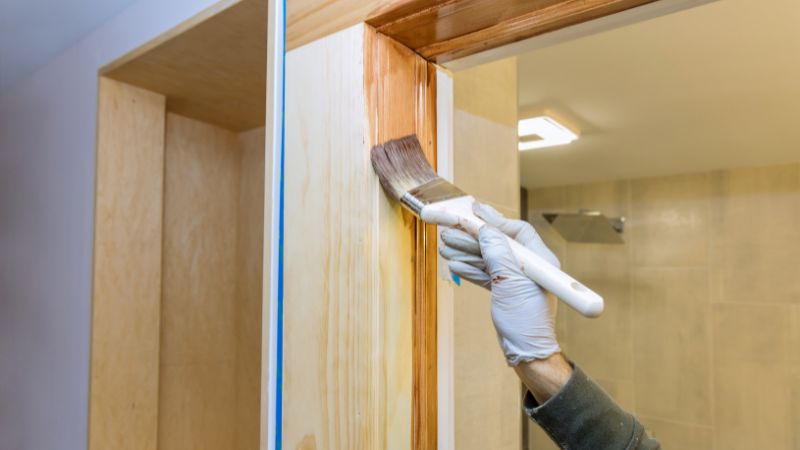 A hand wearing a white glove applying varnish to an interior door frame with a paintbrush, with protective tape lining the edges to ensure a clean finish.
