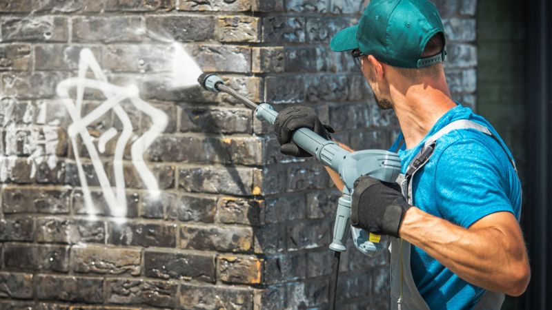 Worker using a high-pressure washer to remove graffiti from a brick wall, preparing the surface for Exterior Painting.