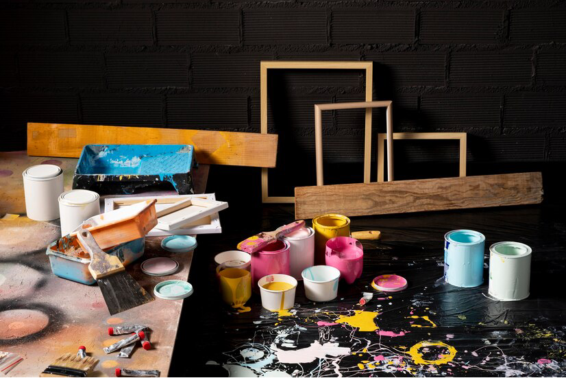 An artist's chaotic work area strewn with vibrant spatters of paint, open cans, mugs daubed with colors, brushes, and unfinished canvases against a backdrop awaiting drywall repair.