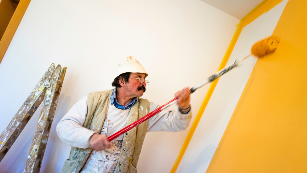 A painting company worker energetically applying a fresh coat of yellow paint on a wall, with his tools of the trade nearby.