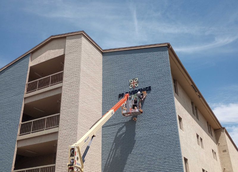 A worker on a telescopic boom lift performing stucco repair on the exterior of a multi-story residential building under a clear blue sky.