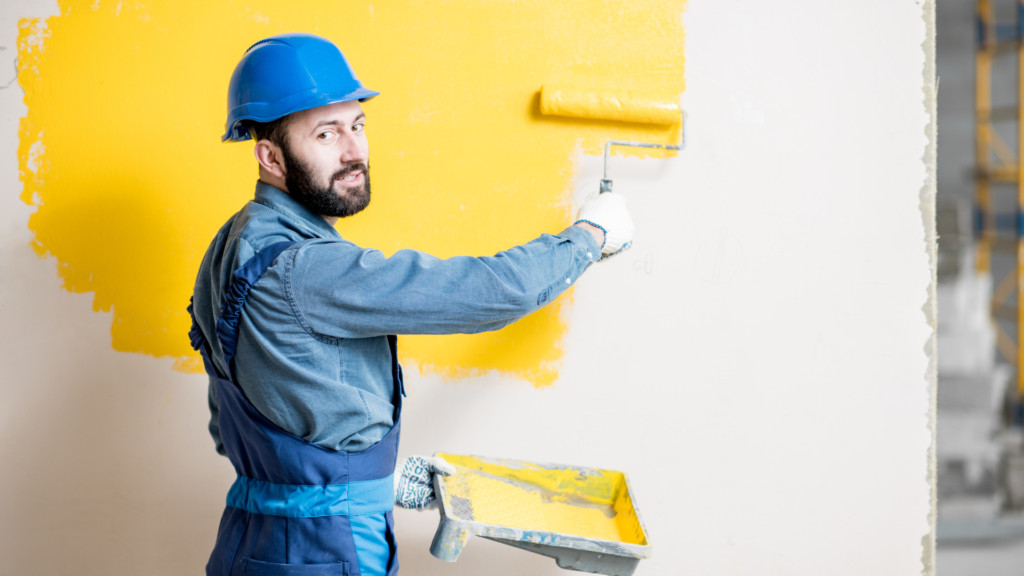 A worker from a painting company in a blue hard hat and coveralls applies bright yellow paint to a wall with a roller.