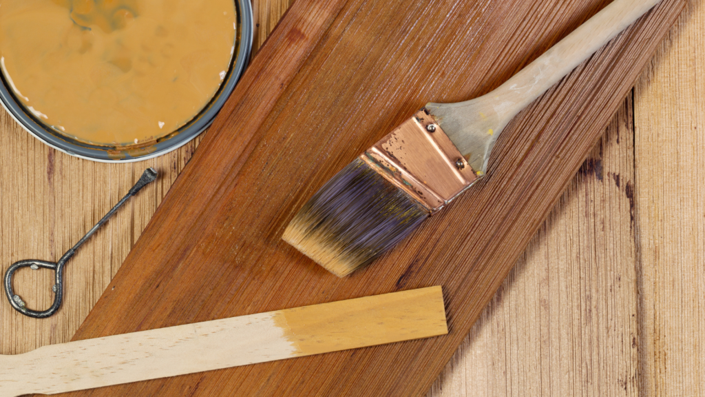 Paintbrush on a wooden surface next to a can of open paint and a paint stirrer, with fresh paint applied on the wood by a professional Painting Company.