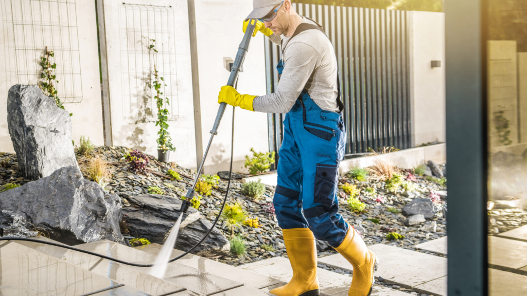 Worker in protective gear using a high-pressure water cleaner outdoors to prepare a surface for stucco repair.