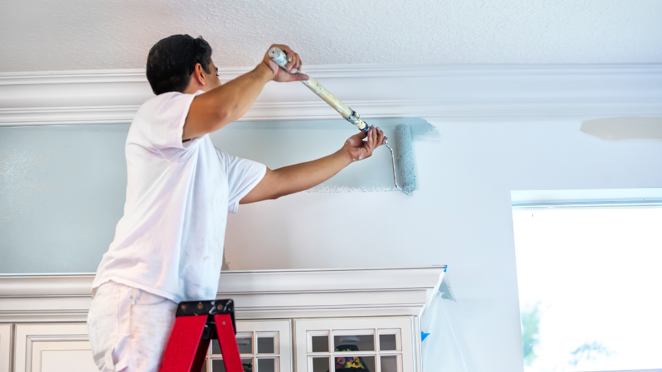 A person on a ladder, part of a professional painting services team, painting the edge of a ceiling with a roller.