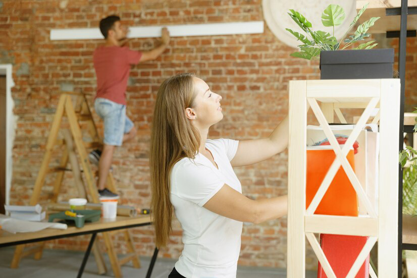 A couple engaging in home improvement, with the woman adjusting items on a shelving unit and the man measuring the wall for interior painting in the background.