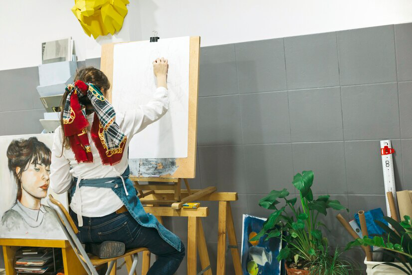 Artist at work in a studio, sketching on a canvas with a vibrant scarf around the neck, surrounded by inspirational elements and exterior painting supplies.