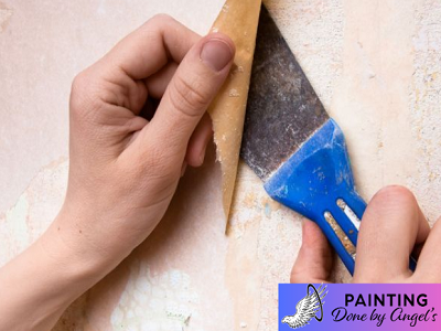 A person's hand using a putty knife to scrape off old wallpaper, with a logo for "Interior Painting by Angel's" in the corner.