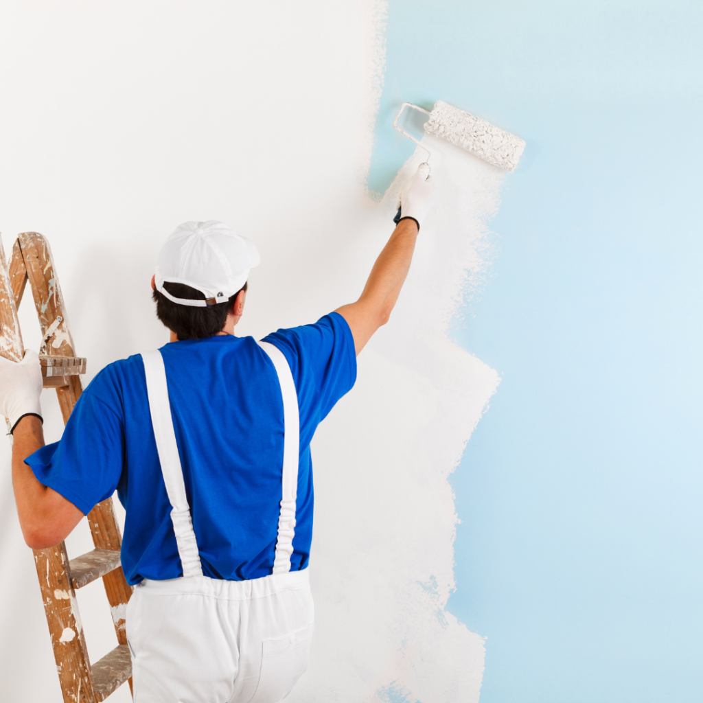 A painter in a blue uniform with a cap and white gloves is using a roller to apply white paint on an exterior wall, standing beside a wooden ladder.