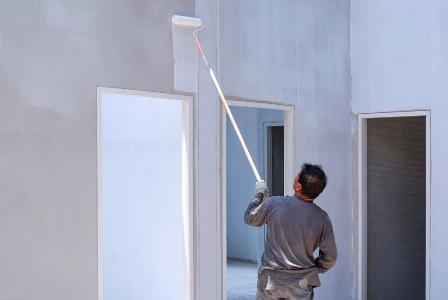 A person from a painting company using a long-handled roller to paint a wall in a room undergoing renovation.