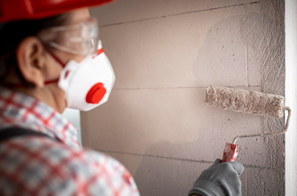 A person wearing safety gear, including a mask and goggles, is performing interior painting by applying paint to a wall with a roller.