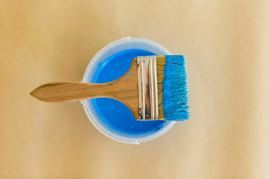 A paintbrush with blue bristles resting on top of an open can of blue paint, viewed from above with a beige background, ready for interior painting.