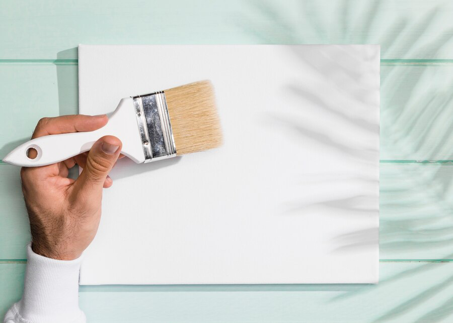A hand holding a paintbrush with white paint over a blank canvas set against a mint green wooden background, ready for interior painting.