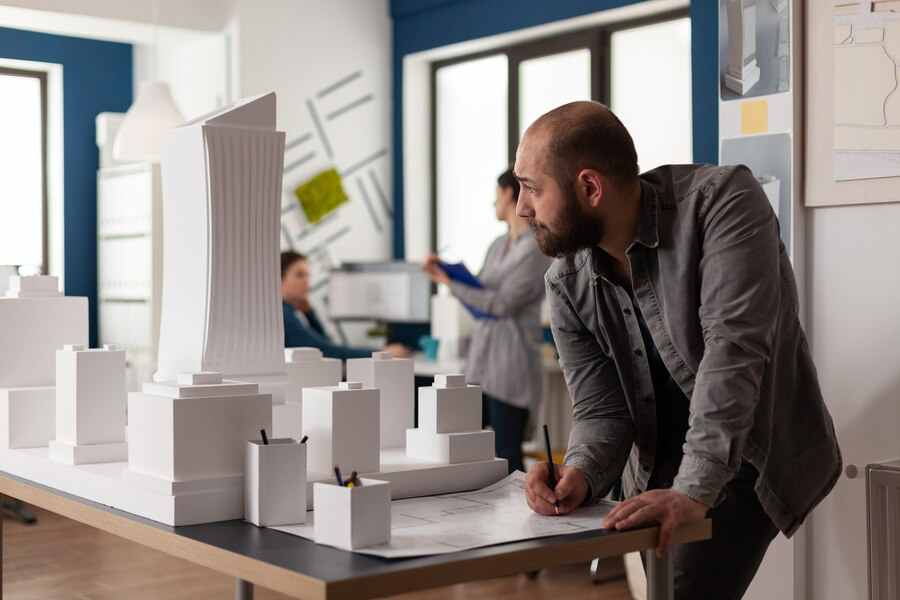 An architect examining a detailed scale model and making notes, with colleagues discussing interior painting plans in the background in a modern office setting.