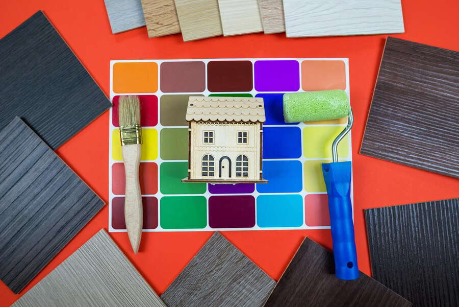 A variety of colorful paint swatches and home improvement material samples, including exterior painting and stucco repair options, surround a small model house, with a paintbrush and roller poised for a creative renovation