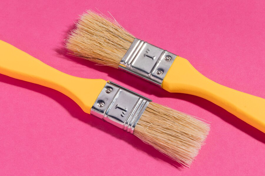 Two yellow paintbrushes with wooden bristles, belonging to a painting company, lying side by side on a vibrant pink background.