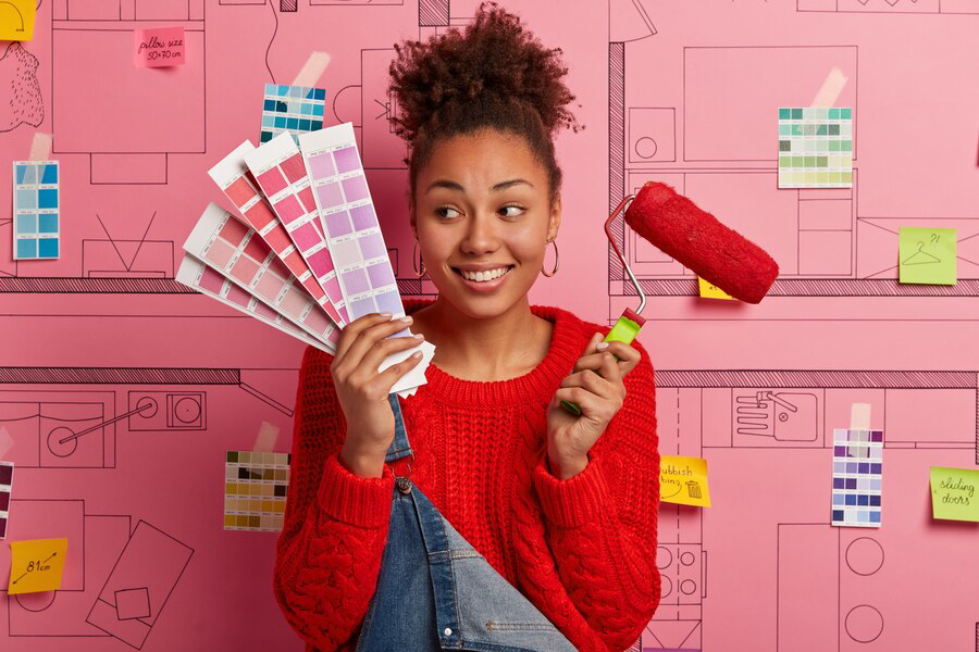 A young woman holding a paint roller and color swatches with a playful smile, surrounded by creative wall sketches and planning notes for home redecoration, ready to embark on exterior painting.