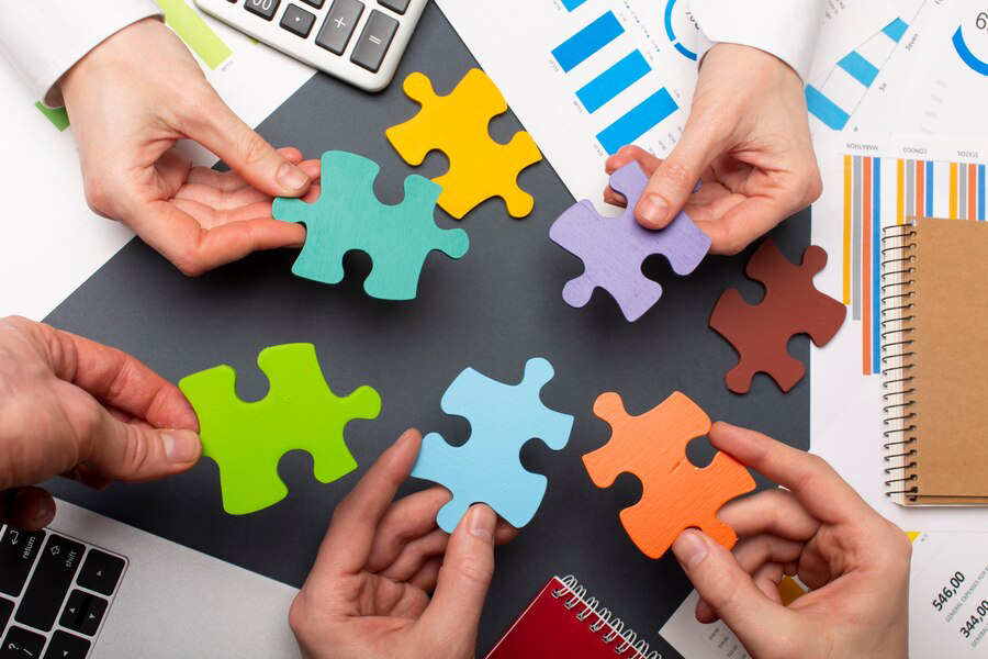 Four hands collaboratively working to fit colorful puzzle pieces together on a desk strewn with business charts, a notebook, and interior painting samples, symbolizing teamwork and problem-solving in a corporate