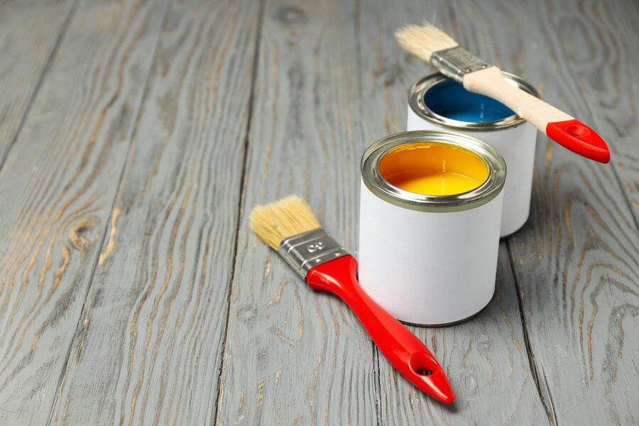 Open paint cans with blue and yellow paint and brushes on a wooden surface, indicating an interior painting project or room renovation in progress.