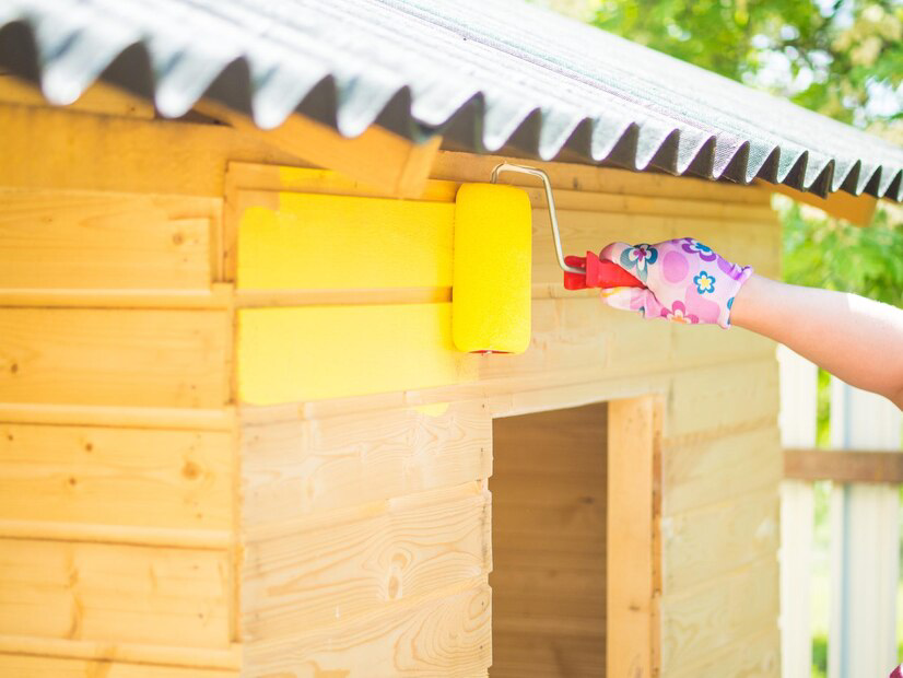 Brightening the playhouse: a vibrant coat of yellow exterior paint is applied to wooden walls under a sunny sky by a professional painting company.