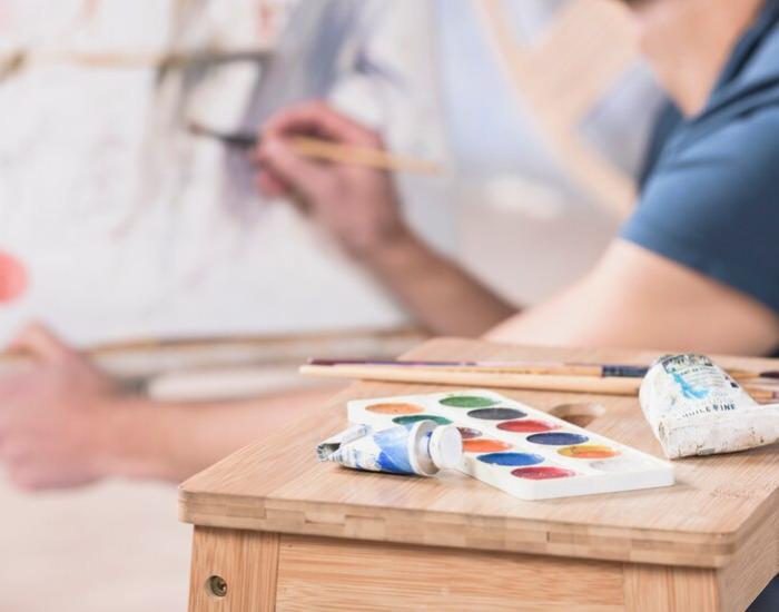 An artist at work behind a wooden easel, with an array of brushes and a colorful palette of watercolors in the foreground, showcasing interior painting techniques.