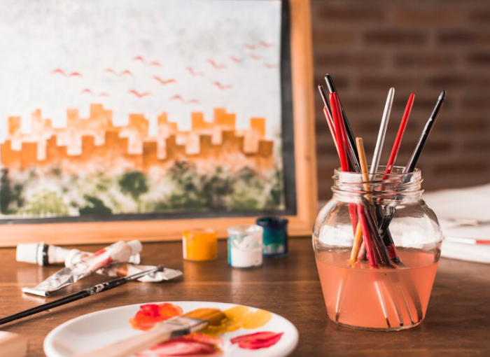A creative workspace with an assortment of paintbrushes in a jar, a palette with fresh dabs of paint, and an inviting canvas with an unfinished cityscape painting that showcases skills in interior painting in